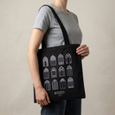 Lifestyle view of a black canvas tote bag carried over a model's shoulder. The front features twelve lavender gothic cathedral window illustrations arranged in a 3-by-4 grid. Centered below the windows, the logo reads ‘MORBID’ in lavender with ‘Live’ in smaller script beneath.
