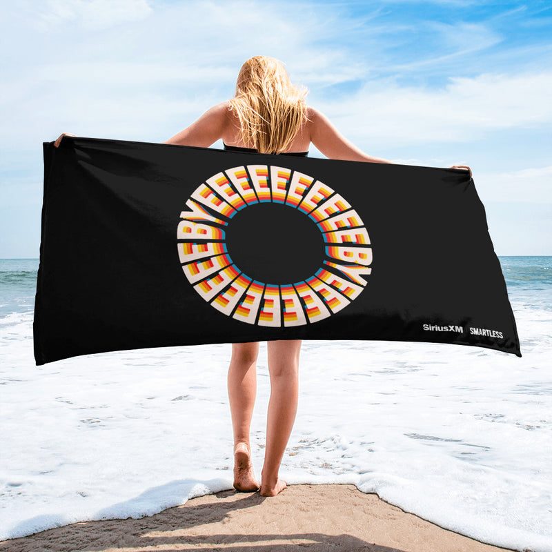 A woman stands on the beach holding a black towel with a bold circular design made of repeating 3D-style 'BYEEEEEEE' text in yellow, orange, and white. In the lower right corner, the towel features the 'SiriusXM' and 'SMARTLESS' logos in white.