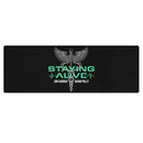 A flat lay of a black yoga mat featuring a centered caduceus — a winged rod with two serpents, representing medicine — above teal text 'STAYING ALIVE' and white names 'JON GABRUS' and 'ADAM PALLY'.