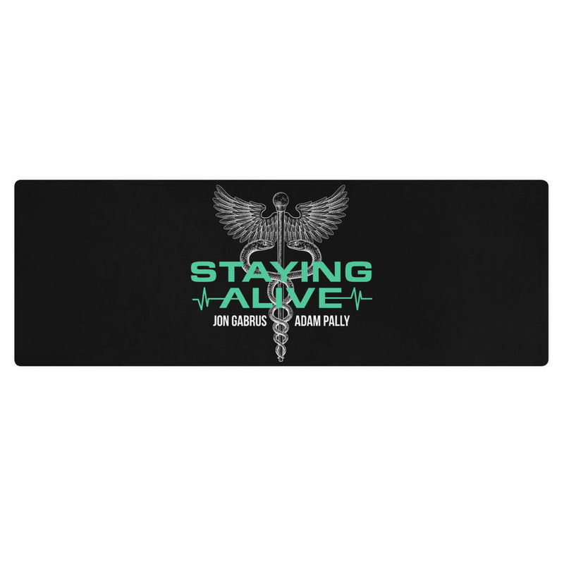 A flat lay of a black yoga mat featuring a centered caduceus — a winged rod with two serpents, representing medicine — above teal text 'STAYING ALIVE' and white names 'JON GABRUS' and 'ADAM PALLY'.