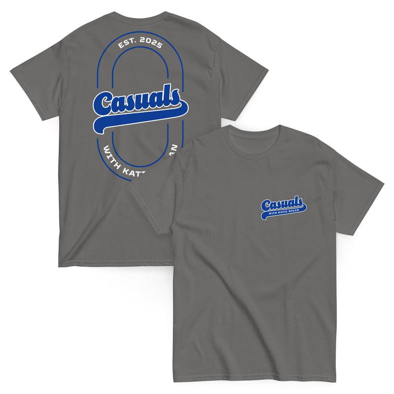 Two charcoal short-sleeve T-shirts laid flat, one showing the front and one showing the back. The front displays a small blue and white 'Casuals WITH KATIE NOLAN' logo on the left chest, and the back features a large blue and white 'Casuals' design with the text 'EST. 2025' and 'WITH KATIE NOLAN' arranged in a circular layout within a double-line oval.