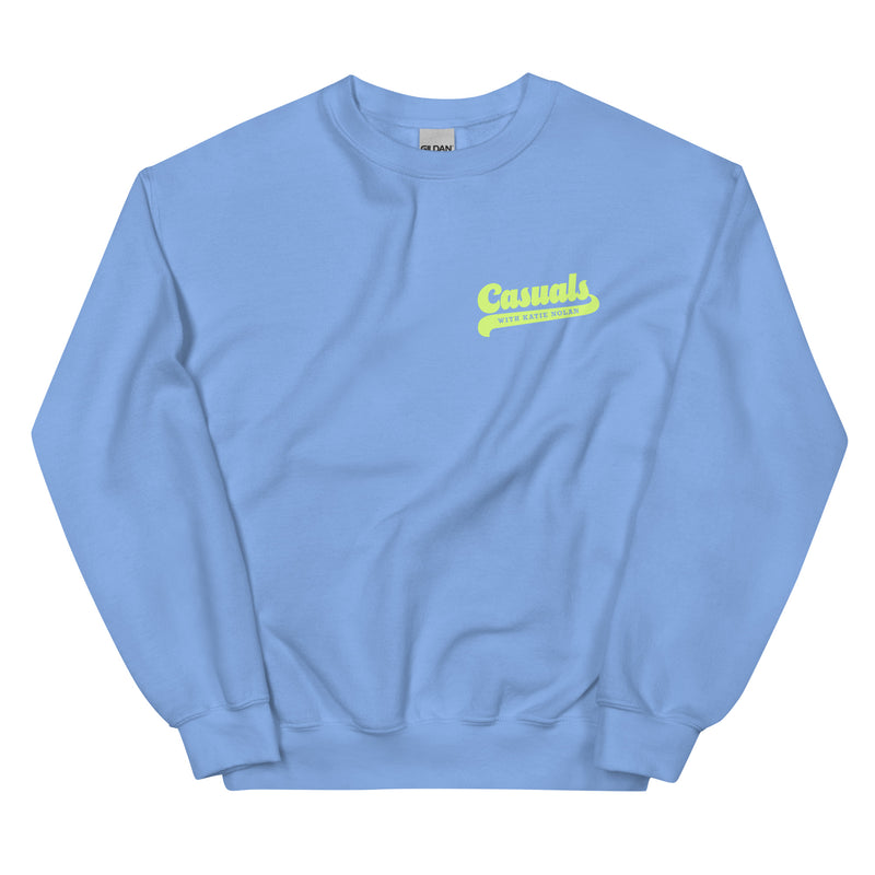 Light blue crewneck sweatshirt laid flat, showing front left chest print of the word 'Casuals' in large yellow script and 'WITH KATIE NOLAN' in smaller all-caps yellow text beneath it.
