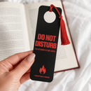 Lifestyle view of a hand holding a black door-hanger-style bookmark in front of an open book. The bookmark has a circular cutout with a red cord and tassel. Red text reads ‘DO NOT DISTURB’ and ‘I’M LISTENING TO VIBE CHECK.’ Near the bottom, a red flame graphic appears above ‘