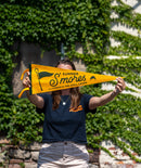 A person holding a yellow pennant that says 'Summer S'mores with Conan & The Chill Chums' in front of a green ivy-covered wall.