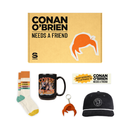 A flat lay showing the ‘CONAN O’BRIEN NEEDS A FRIEND’ Fan Box and it's contents, including the orange box, socks, black mug, bumper sticker, keychain, and black corduroy hat with matching white embroidered logo.