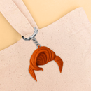 A keychain shaped like a swooped tuft of bright orange hair hangs from a beige canvas tote bag handle against a soft tan background.
