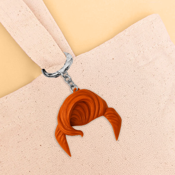 A keychain shaped like a swooped tuft of bright orange hair hangs from a beige canvas tote bag handle against a soft tan background.