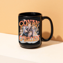A black ceramic mug featuring an orange and white collage design of ‘CONAN O’BRIEN,’ with multiple images of Conan O’Brien in different poses, including holding a guitar and gesturing animatedly. The mug sits on a light-colored surface against a peach background.