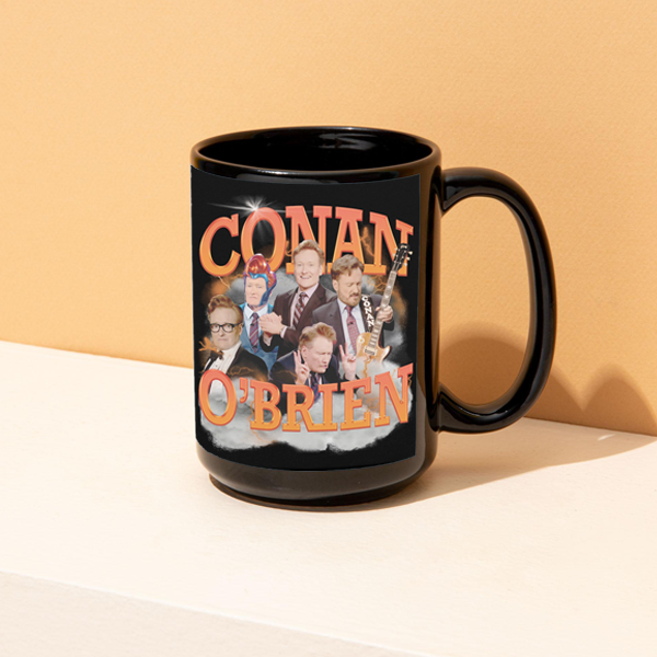 A black ceramic mug featuring an orange and white collage design of ‘CONAN O’BRIEN,’ with multiple images of Conan O’Brien in different poses, including holding a guitar and gesturing animatedly. The mug sits on a light-colored surface against a peach background.