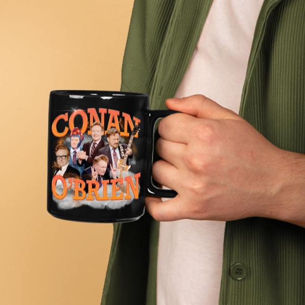 A person wearing a green jacket holds a black mug featuring the orange ‘CONAN O’BRIEN’ collage design, showing Conan O’Brien in various expressions and outfits.