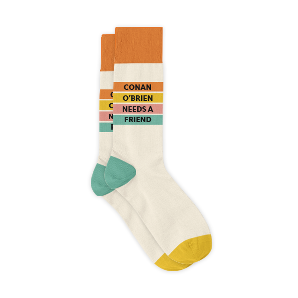 A pair of cream-colored crew socks with colored stripes in orange, yellow, pink, and teal across the top, featuring black text that reads ‘CONAN O’BRIEN NEEDS A FRIEND.’ The socks have mustard yellow toes, teal heels, and orange cuffs.