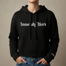Lifestyle view of a black cropped hoodie worn by a model. The front features large white gothic blackletter text reading ‘Immortally Weird’ across the chest. The hoodie has a drawstring hood, cropped fit, long sleeves, and a small sleeve print near the wearer’s left cuff reading ‘MORBID Live’.