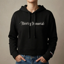 Lifestyle view of a black cropped hoodie worn by a model. The front features large white gothic blackletter text reading ‘Weirdly Immortal’ across the chest. The hoodie has a drawstring hood, cropped fit, long sleeves, and a small print near the wearer’s left cuff reading ‘MORBID Live’.