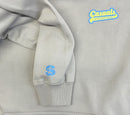 Detail view of an off-white hoodie with blue and yellow ‘Casuals with Katie Nolan’ logo printed on the left chest and a small blue SiriusXM S-Star logo on the left wrist cuff.