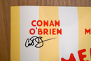 Close-up of the 'Conan O'Brien' section of the 'Summer S’mores with Conan & The Chill Chums' poster, featuring his name in red above a bold black signature extending across the yellow stripe.