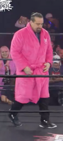 Wrestler in a pink robe walking toward the ring with fans in the background, some wearing pink clothing.