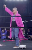 Man in a pink robe holding a microphone in a wrestling ring, with fans cheering in the background.