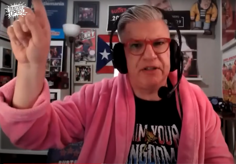 Person wearing a pink robe and headset speaking animatedly in a decorated room filled with memorabilia and posters.
