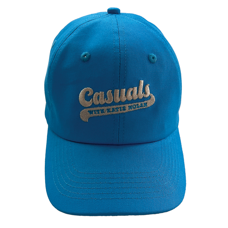 Straight-on view of a bright blue baseball cap featuring beige embroidery of the ‘Casuals with Katie Nolan’ logo centered on the front panel.