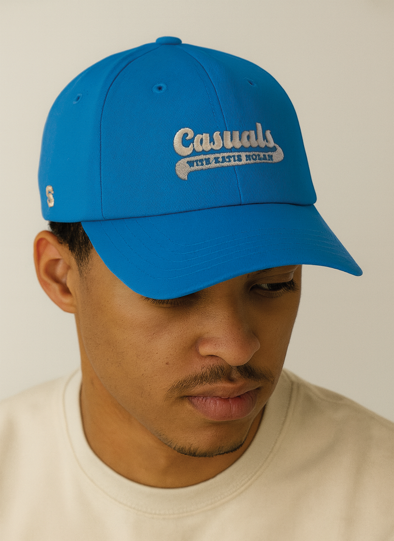 A person wearing a bright blue baseball cap with the beige embroidered ‘Casuals with Katie Nolan’ logo on the front and the ‘S-Star’ SiriusXM logo on the left side, paired with a cream-colored shirt against a neutral background.