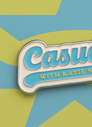 Close-up of the 'Casuals with Katie Nolan' enamel pin showing the glossy blue enamel, gold lettering, and silver metal edges, placed on a yellow and blue star-patterned card background.