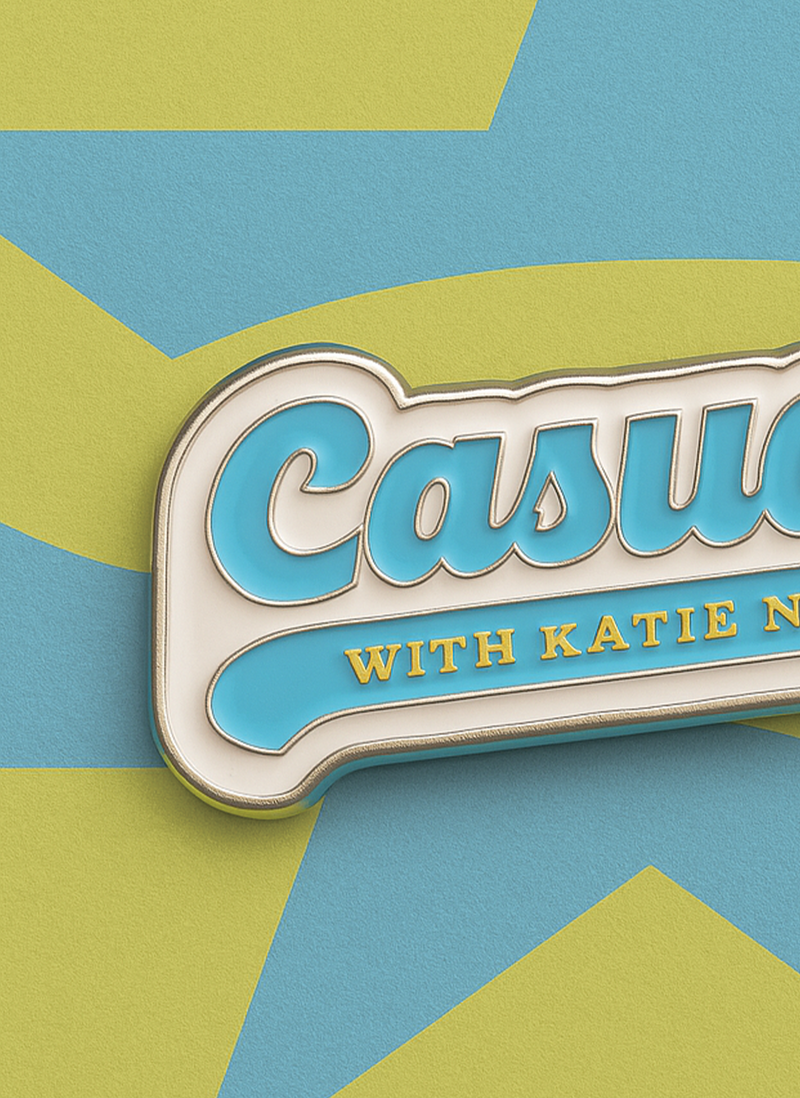 Close-up of the 'Casuals with Katie Nolan' enamel pin showing the glossy blue enamel, gold lettering, and silver metal edges, placed on a yellow and blue star-patterned card background.