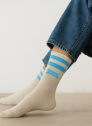 Close-up of a person wearing cream-colored ribbed crew socks with two sky-blue stripes around the cuffs and the word ‘Casuals’ in matching blue text above the ankle. The person is seated indoors on a light floor, wearing cropped blue jeans.