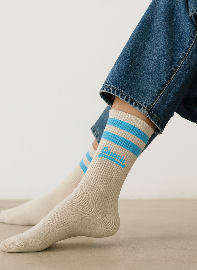 Close-up of a person wearing cream-colored ribbed crew socks with two sky-blue stripes around the cuffs and the word ‘Casuals’ in matching blue text above the ankle. The person is seated indoors on a light floor, wearing cropped blue jeans.