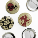 Flat lay of a 3-piece pin button set on a light background, showing three illustrated designs and two pin-back views. Designs include overlapping skulls—one deep red and one bone-white—on a distressed beige background; a repeating black-and-cream pattern of startled vintage-style faces; and a red winged devil-like figure with pointed ears and tail on a speckled beige background. Metal pin backs are visible on two buttons.