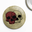Close-up of a single round pin button featuring two overlapping skull illustrations, one dark red in front and one off-white behind, both with hollow eye sockets and detailed teeth, set against a textured beige background with a worn, vintage look.
