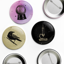Flat lay of a 3-piece pin button set showing multiple designs and pin-back views. Visible designs include a lavender button with a crystal ball illustration, a beige button with a black crow perched on a branch, and a black button featuring a lit candle with wax dripping into a snake below; metal pin backs are shown on two buttons.
