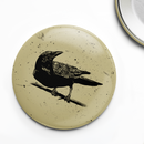 Close-up of a round pin button featuring a detailed black illustration of a crow perched on a branch, set against a distressed beige background with scattered speckled texture for a vintage, worn effect.