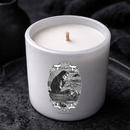 Close-up angled view of a white candle in a glass jar with a centered wick, featuring a detailed black illustration of a hooded skeleton rowing a boat. The candle sits on a dark textured surface with a moody, shadowy background.