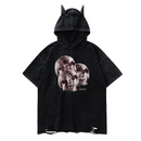 Front view of a black washed short-sleeve hoodie with a pointed hood featuring two small horn-like protrusions. The chest displays three overlapping realistic skull illustrations in muted red tones with small ‘MORBID’ text beneath. The hem has distressed, frayed holes for a worn, edgy look.