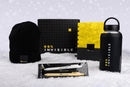 A landscape holiday flat lay featuring 99% Invisible merchandise arranged on white snow. From left to right: a black ribbed beanie with a small rectangular patch reading ‘99% Invisible’ in white and yellow text; a black and yellow grid-patterned gift box with the ‘99% Invisible’ logo printed in white and yellow on the lid; a yellow and black spiral notebook with a geometric cover design and the ‘99% Invisible’ logo printed near the bottom; a matte black water bottle with the ‘99% Invisible’ logo printed ver