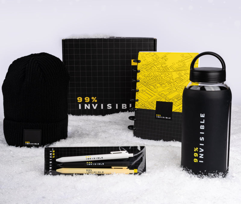 A holiday-themed flat lay of 99% Invisible merchandise displayed on white snow. Items include a black ribbed beanie, a black and yellow grid-patterned gift box, a yellow and black spiral notebook, a matte black water bottle printed with ‘99% INVISIBLE’ in white and yellow text, and a three-pen set in white, black, and tan packaging.