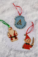 Three enamel ornaments—one blue and gold with text reading ‘COMEDY BANG! BANG!’ around a biker illustration, one of Santa Claus making a rude hand gesture, and one of a mischievous elf holding a knife—arranged on a snowy background.