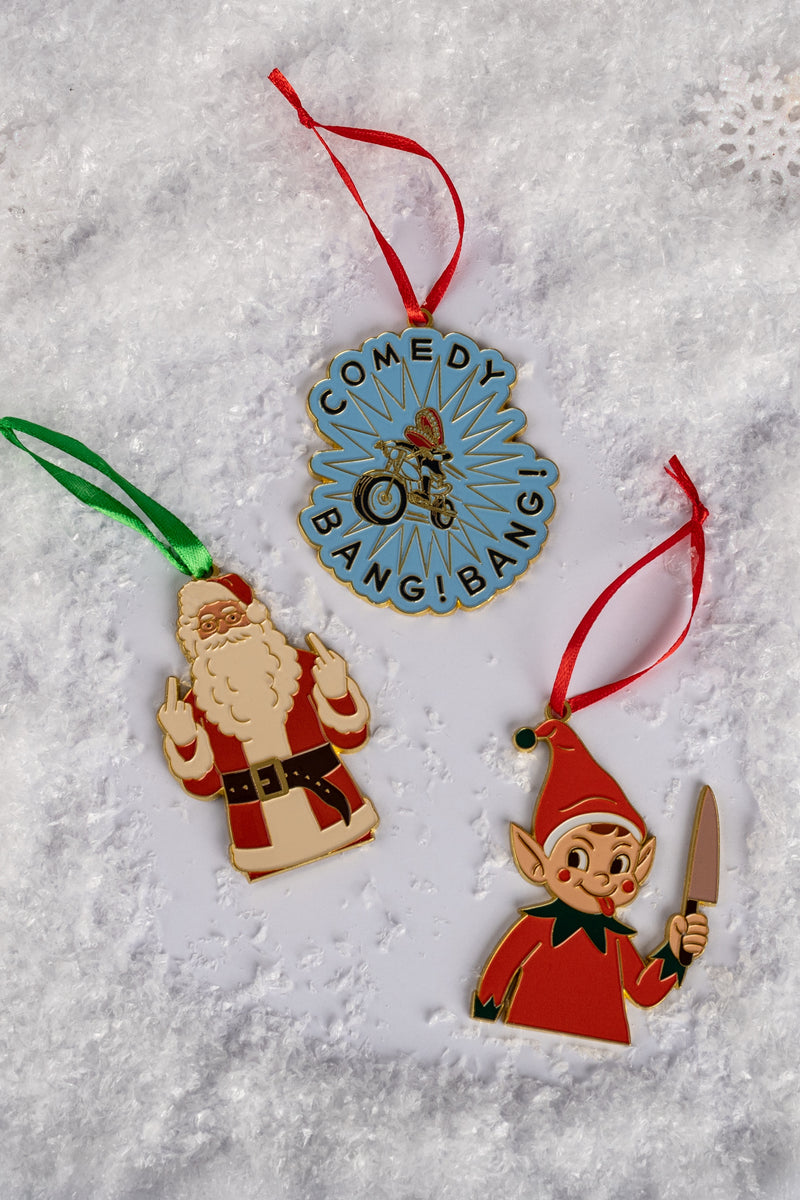 Three enamel ornaments—one blue and gold with text reading ‘COMEDY BANG! BANG!’ around a biker illustration, one of Santa Claus making a rude hand gesture, and one of a mischievous elf holding a knife—arranged on a snowy background.