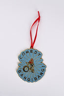 Blue ornament with a jagged edge design featuring the text ‘COMEDY BANG! BANG!’ in black around an illustration of a person riding a bike, hanging from a red ribbon.