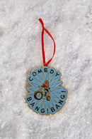 A blue enamel ornament featuring the phrase ‘COMEDY BANG! BANG!’ encircling an illustration of a biker wearing a red helmet, shown on snow-like textured ground.