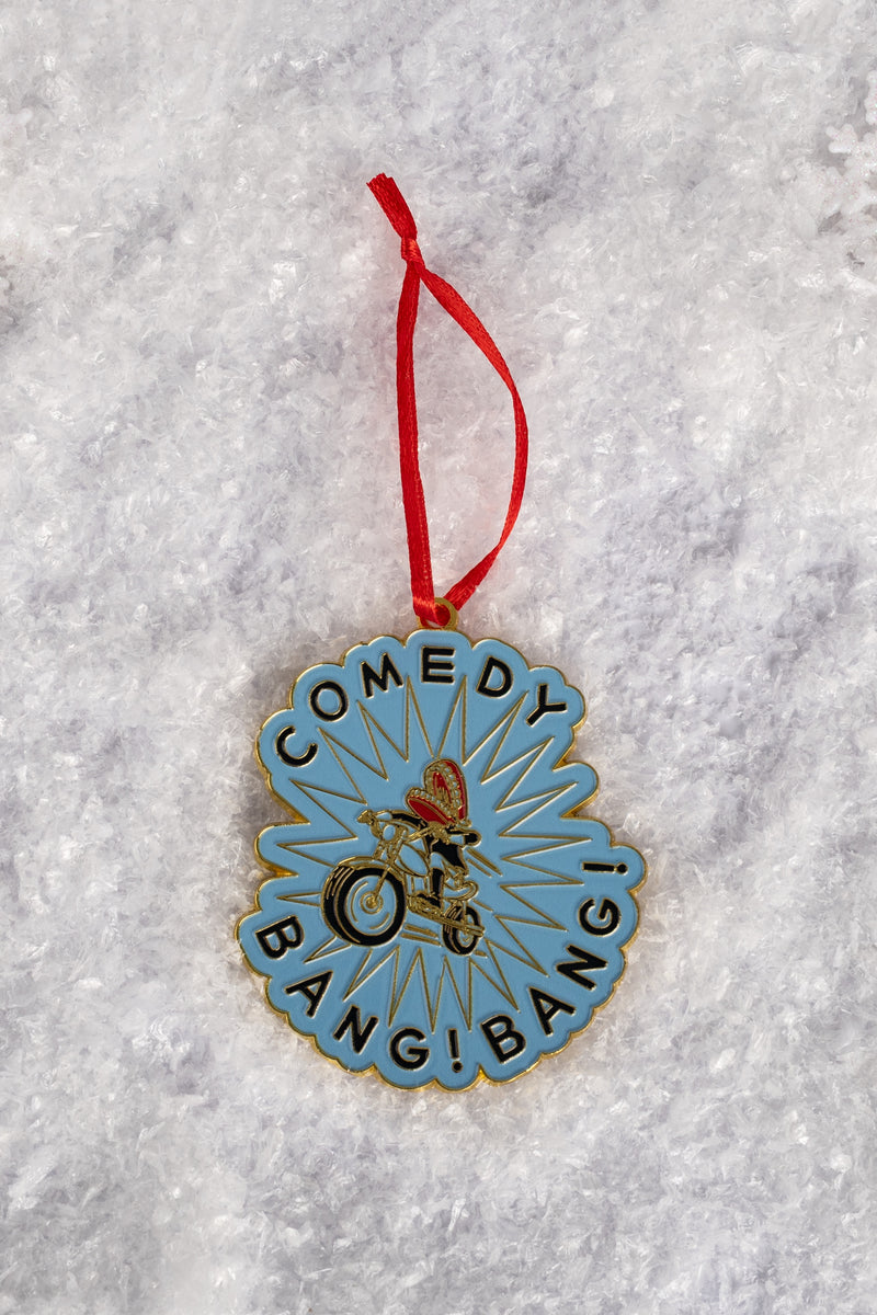 A blue enamel ornament featuring the phrase ‘COMEDY BANG! BANG!’ encircling an illustration of a biker wearing a red helmet, shown on snow-like textured ground.