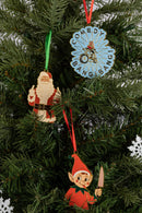 Three enamel ornaments hanging on a Christmas tree: a Santa ornament with a green ribbon, a blue ‘COMEDY BANG! BANG!’ ornament with a red ribbon, and an elf ornament with a red ribbon and a knife.