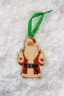 A close-up of the Santa ornament featuring Santa Claus in red and white attire, holding up both middle fingers, hanging from a green ribbon against a snowy background.