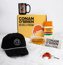 A holiday-themed flat lay of ‘Conan O’Brien Needs a Friend’ merchandise arranged on snow. A yellow branded box with the podcast’s logo sits in the center with a black mug on top featuring multiple images of Conan O’Brien and the same logo in orange text. In front are a black corduroy cap embroidered with a white outline of Conan’s hair, a yellow and orange striped pair of socks reading ‘CONAN O’BRIEN NEEDS A FRIEND’, a rectangular bumper sticker with the phrase ‘KEEP HONKING!! I’M LISTENING TO CONAN O’BRIEN