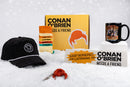 A landscape version of the holiday flat lay featuring ‘Conan O’Brien Needs a Friend’ merchandise displayed on snow. Centered is the yellow podcast box with Conan’s hair graphic, flanked by a black mug with Conan’s image on the right and socks, hat, bumper sticker, and red hair keychain arranged neatly in front. The white background and snow texture create a festive, winter promotional display.