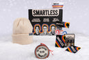 Snow-covered display of the ‘SMARTLESS’ holiday gift set. A black box with ‘SMARTLESS’ text above hosts ‘Jason Bateman,’ ‘Sean Hayes,’ and ‘Will Arnett’ is surrounded by a cream beanie, rainbow-striped bowl, black sticker with ‘SMARTLESS,’ die-cut host stickers, and a round ornament that reads ‘'TIS THE SEASON TO BE SMARTLESS.’