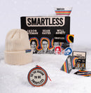 Snowy flat lay of the ‘SMARTLESS’ podcast gift set. A black box with ‘SMARTLESS’ text and images of hosts ‘Jason Bateman,’ ‘Sean Hayes,’ and ‘Will Arnett’ is surrounded by a cream beanie with a blue ‘SMARTLESS’ tag, a white bowl with rainbow stripes, a sticker, and a round ornament reading ‘'TIS THE SEASON TO BE SMARTLESS.’