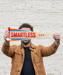 Person holding a pennant flag with 'SMARTLESS' in red text surrounded by stripes in blue, red, orange, and yellow.