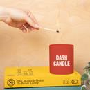 A red candle jar with white text reading ‘DASH CANDLE,’ resting on a yellow book as a hand reaches in to light the wick with a match.