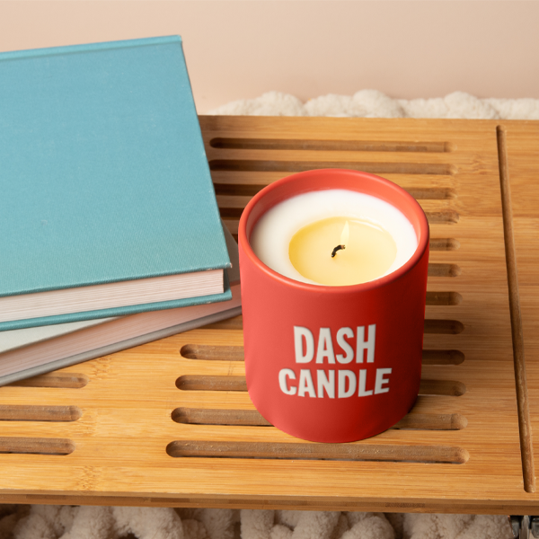 A red candle jar with white text that reads ‘DASH CANDLE,’ placed on a wooden slatted surface beside two closed blue books.